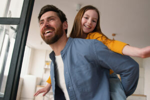 Smiling father carrying child at home for Daddy Do-Over plastic surgery information