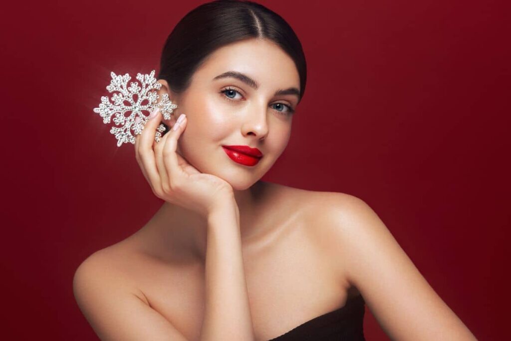 Woman with flawless makeup holding sparkling diamond accessory against red background for holiday beauty inspiration.
